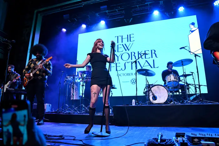 A singer performs on stage in front of a blue screen that says "New Yorker festival."